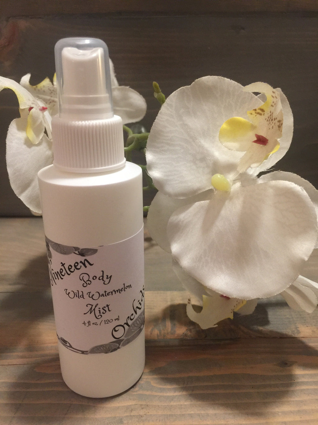 May include: A white bottle with a clear spray nozzle. The bottle is labeled "Nineteen Body Wild Watermelon Mist" and "4 fl oz / 120 ml". The label also has a floral design with the word "Orchid" written on it. The bottle is sitting on a wooden surface with a white orchid flower behind it.